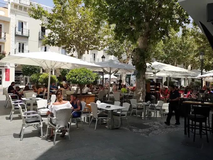 Terraza con sombrillas blancas bajo los árboles de la plaza, llena de clientes