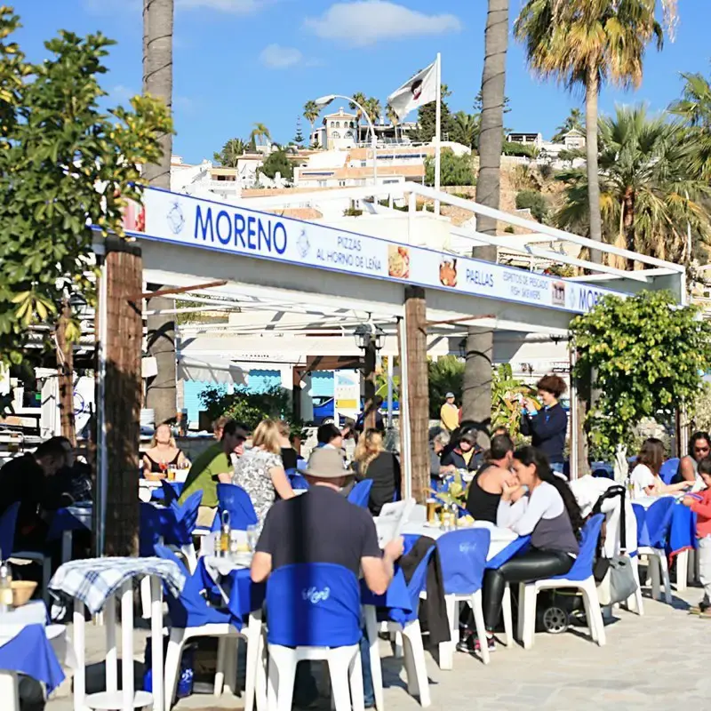 Terraza con sillas azules bajo el toldo que anuncia pizzas al horno de leña y paellas