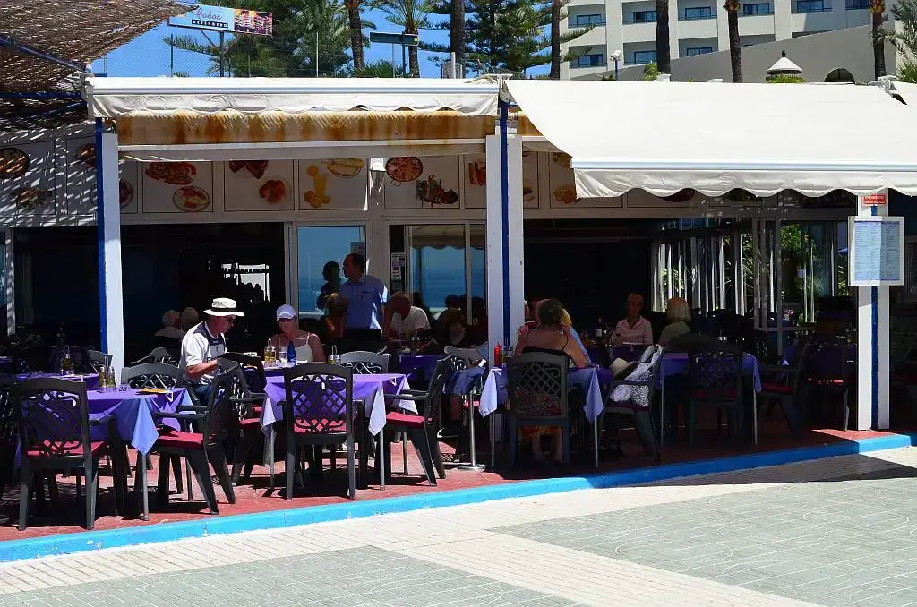 Fachada con toldos blancos y mesas con manteles morados junto al paseo marítimo