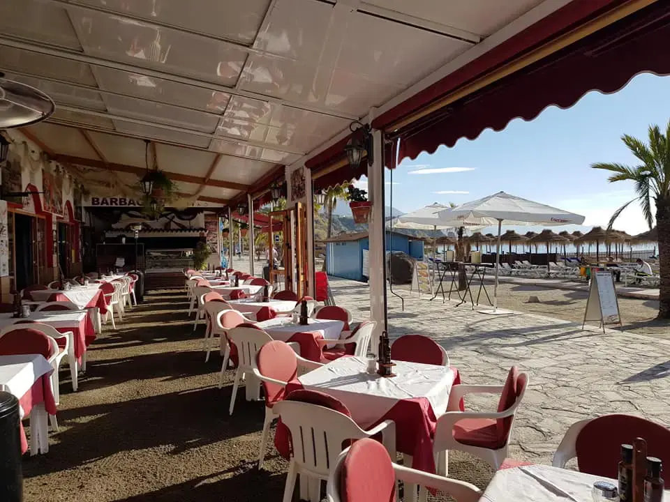 Terraza del Merendero José Cruz con manteles rojos, abierta a la playa y sus sombrillas