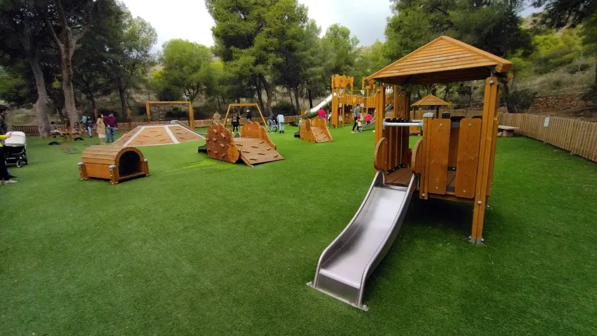 Casita de madera con tobogán metálico y túneles en el parque infantil, rodeado de pinos