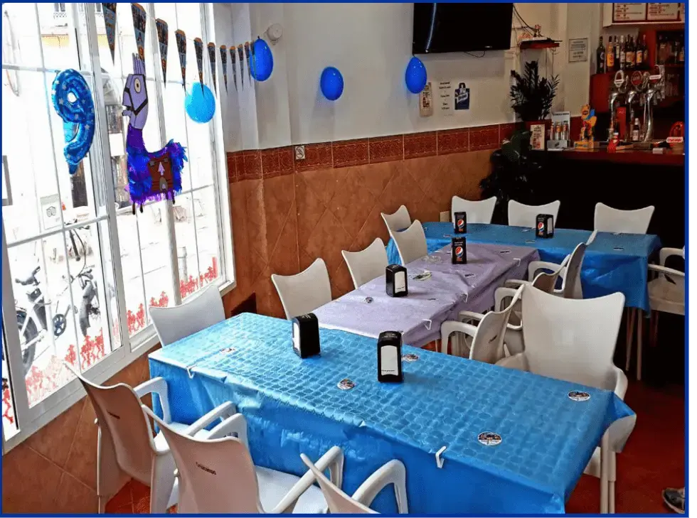 Comedor con manteles azules, sillas blancas, globos y barra con grifos al fondo