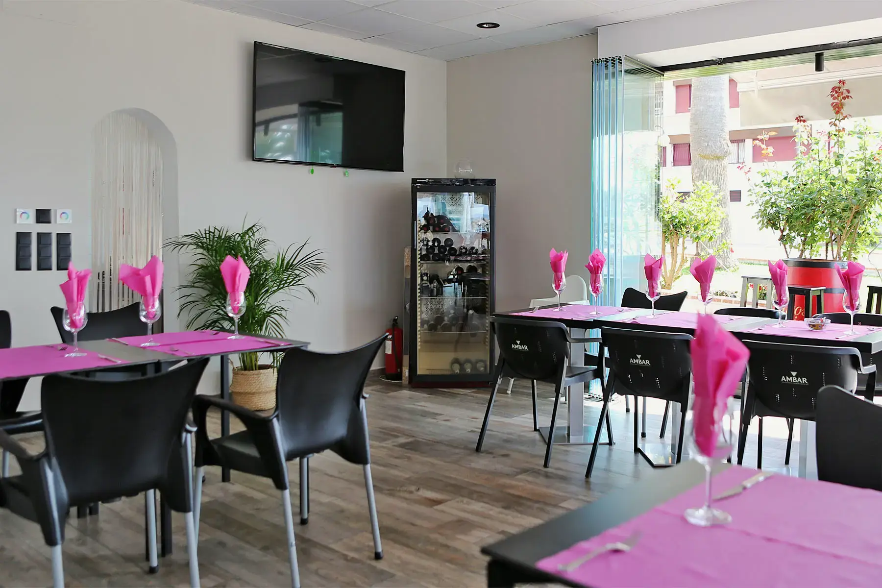 Comedor luminoso con servilletas rosas en las copas, sillas negras y televisión