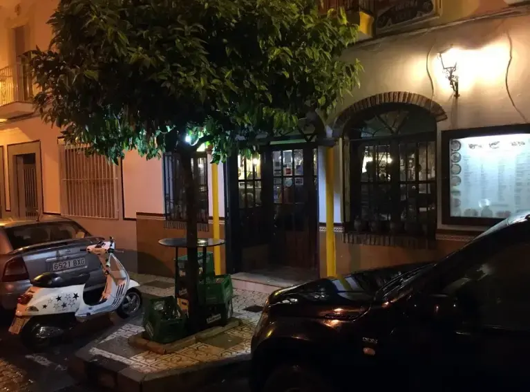 Entrada de la taberna de noche, con naranjo, farol encendido y carta expuesta