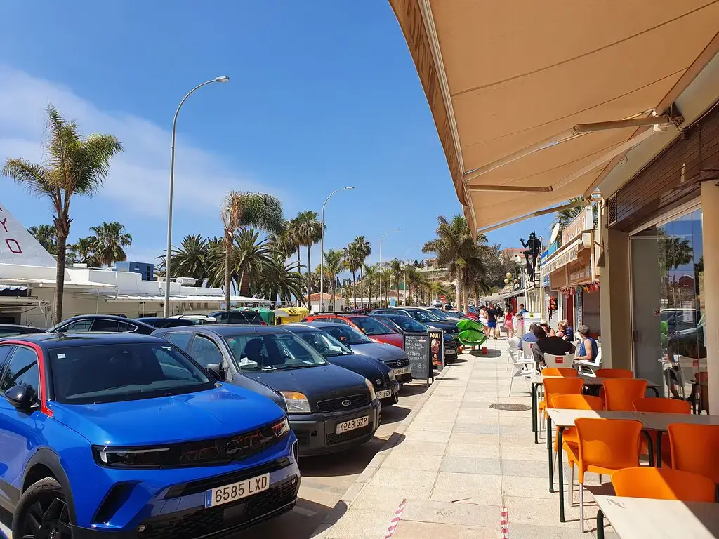 Paseo con palmeras y coches aparcados junto a la terraza de sillas naranjas