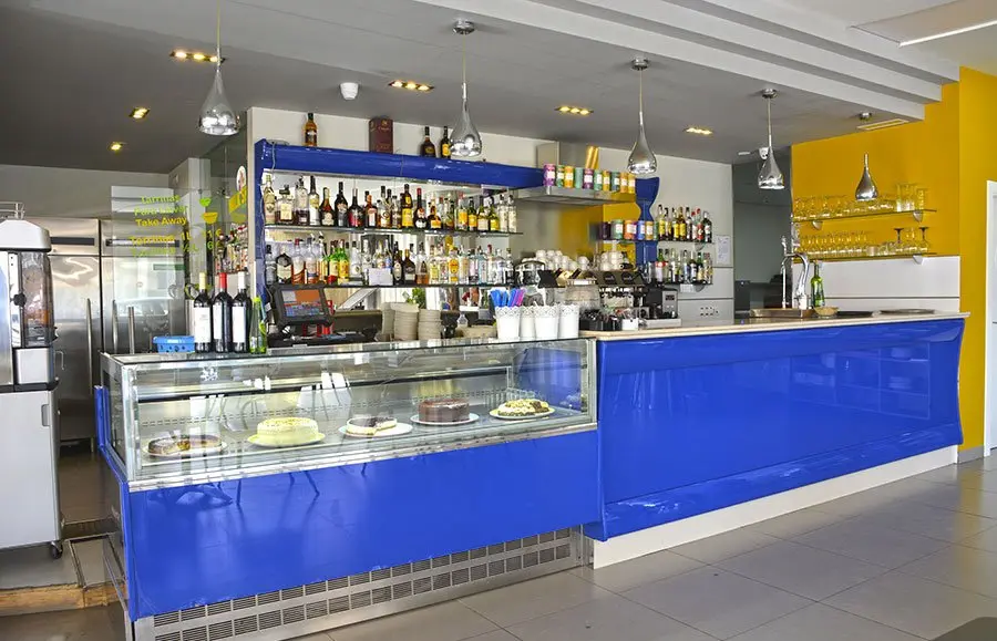 Barra azul con vitrina de tartas y estanterías de bebidas en la Cafetería Albi