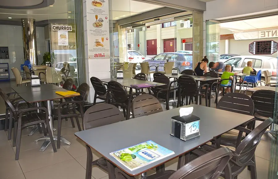 Mesas interiores junto a la cristalera abierta, con cartel de churros y clientes desayunando