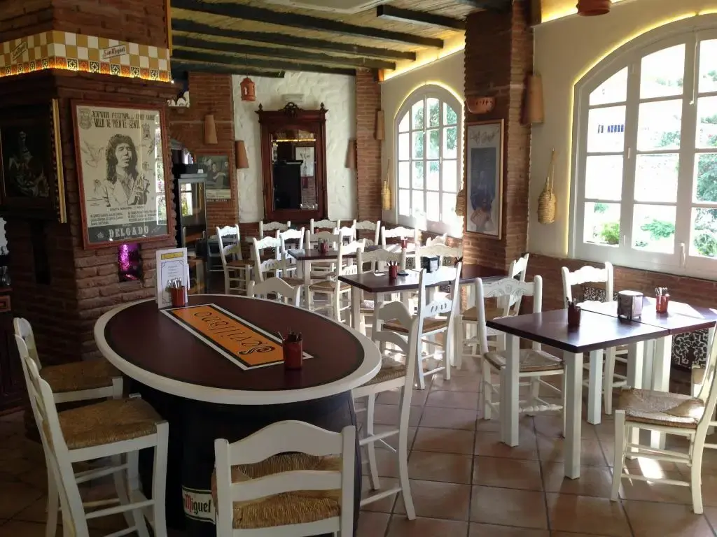 Comedor con sillas blancas de enea, ventanales en arco y un cartel antiguo de flamenco