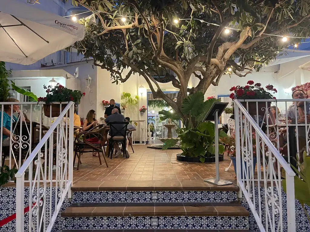 Patio iluminado de noche con mesas bajo un árbol, guirnaldas de luces y escalera de azulejos
