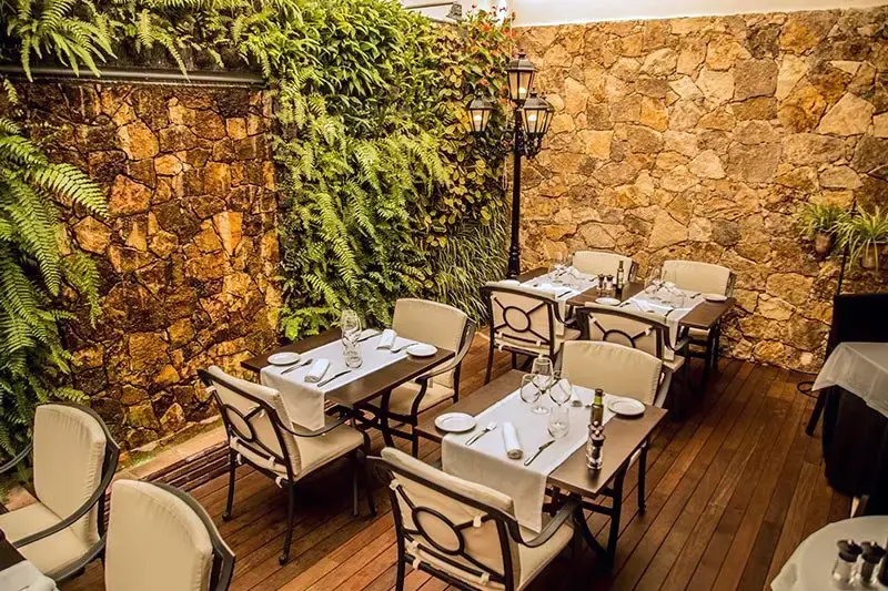 Mesas preparadas sobre tarima de madera junto a helechos, muro de piedra y farola