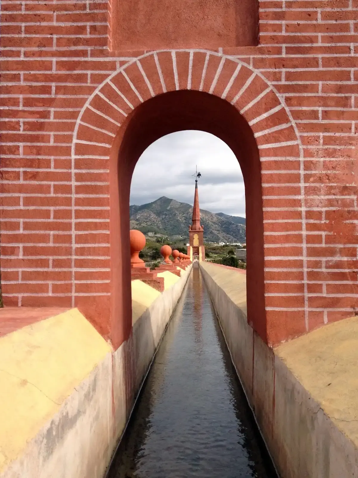 Canal del acueducto visto a través de un arco de ladrillo, con la torrecilla y la sierra al fondo