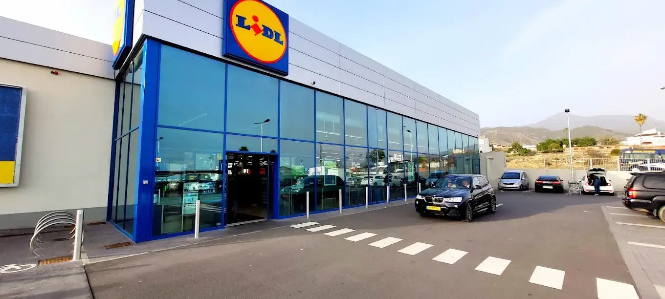 Fachada azul acristalada de Lidl Nerja con coches en el aparcamiento y la sierra al fondo