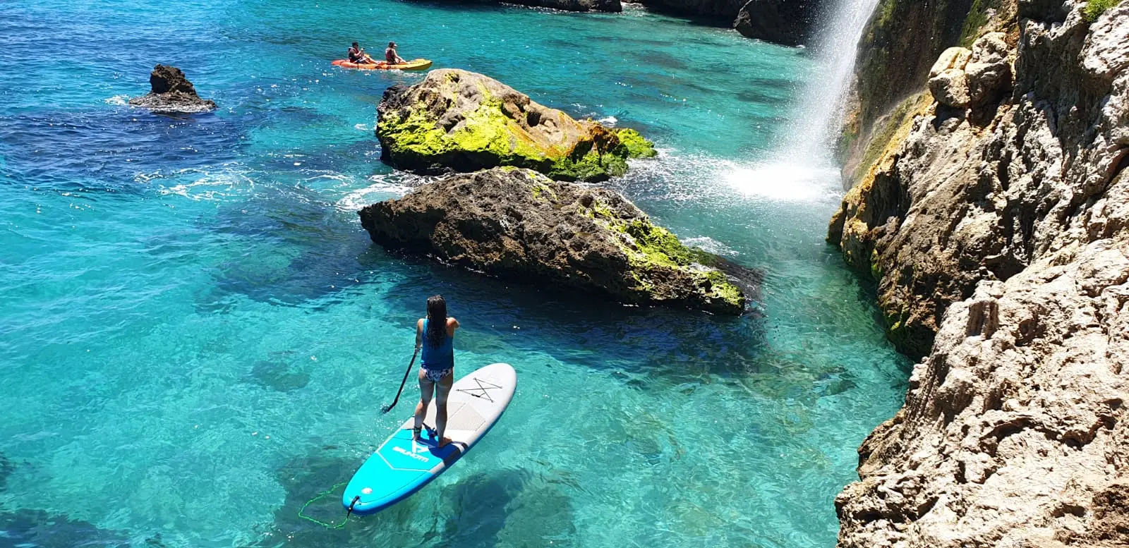 Paddle surf en Maro junto a la cascada y las rocas con musgo, con kayaks al fondo