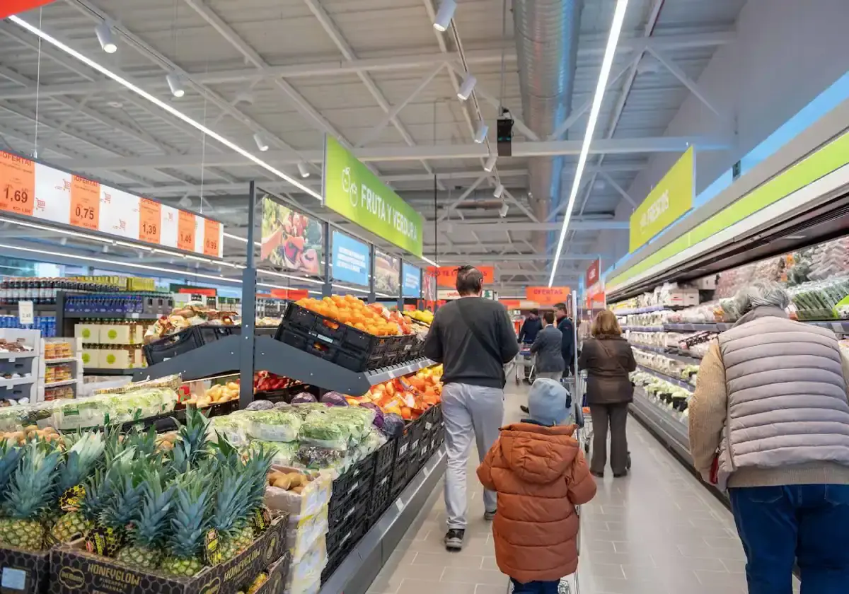 Clientes comprando en el pasillo de fruta y verdura con piñas y precios en lo alto