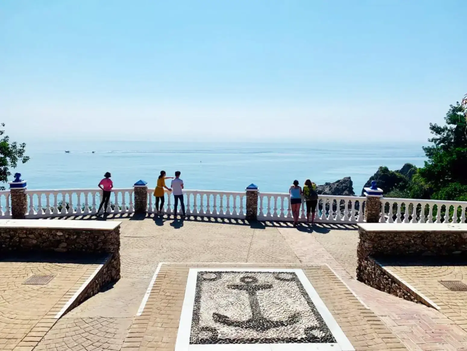 Ancla de cantos rodados en el suelo del mirador y gente asomada a la balaustrada sobre el mar