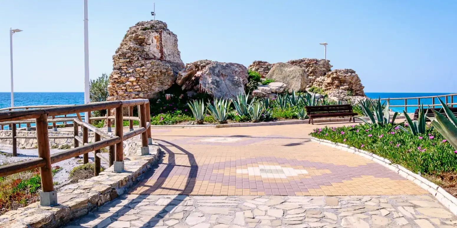 Pasarela de madera hacia las rocas de la antigua torre, con pitas y un banco frente al mar