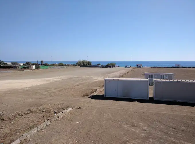 Explanada de tierra del parking El Playazo con casetas prefabricadas y el mar al fondo