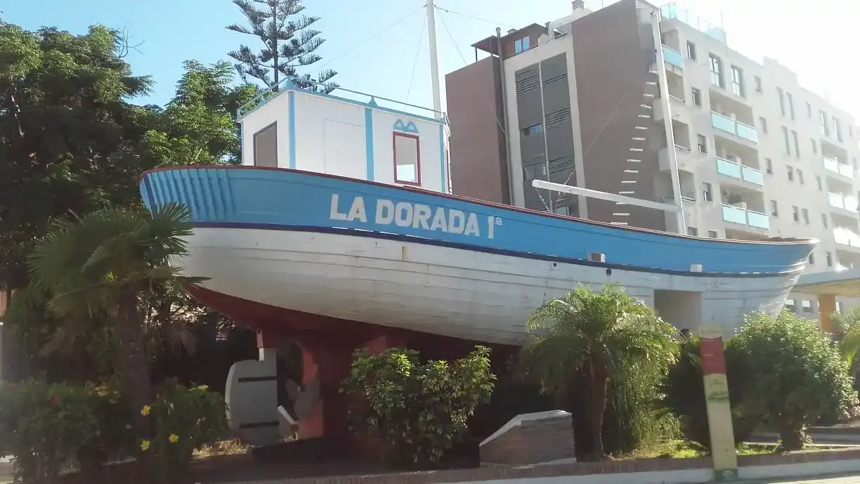 Costado del Barco de Chanquete con su nombre pintado, entre palmeras y edificios