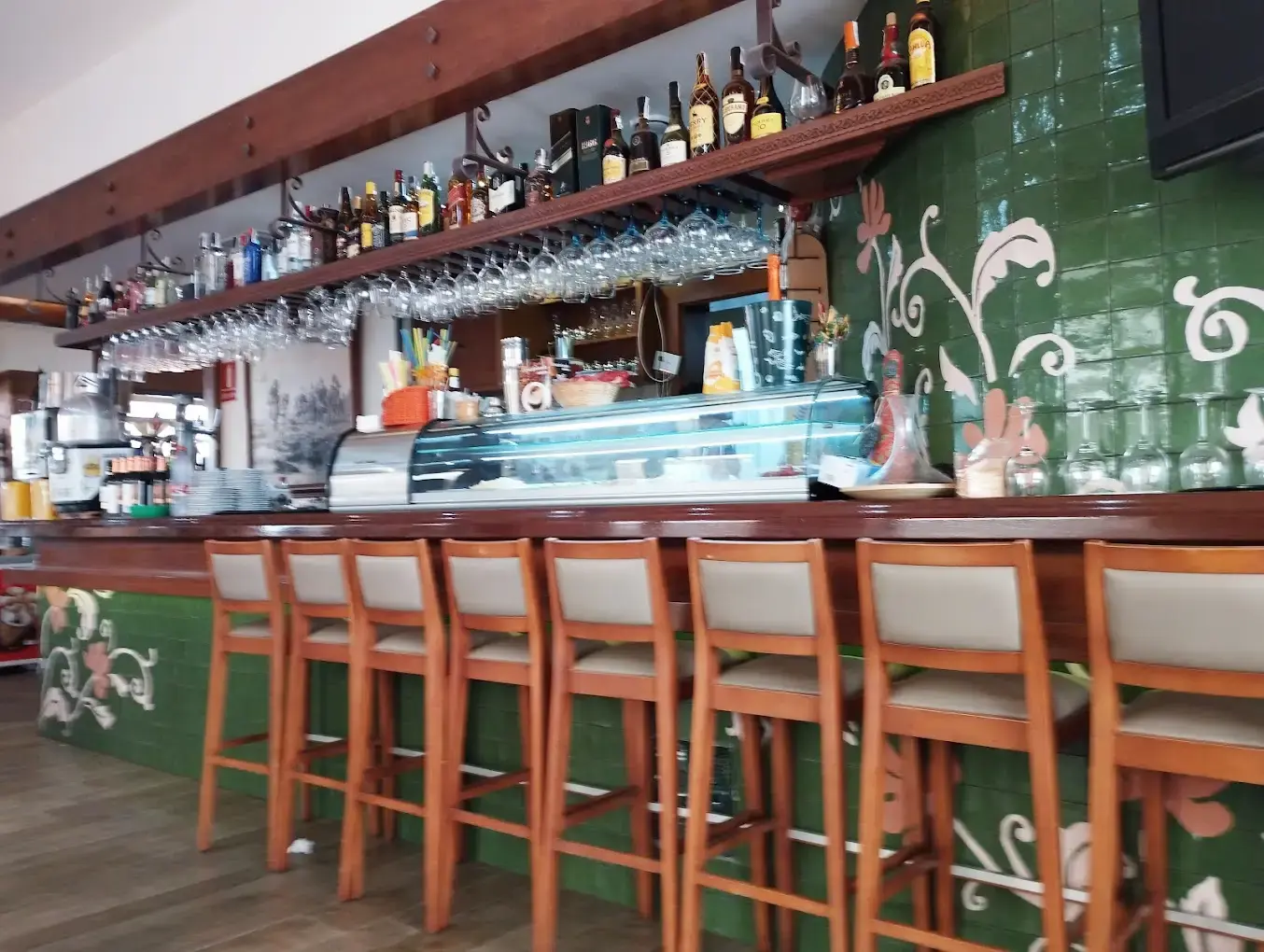 Barra de azulejos verdes con taburetes de madera, vitrina de tapas y botellero