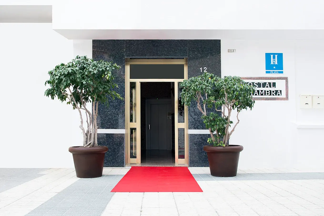 Entrada del Hostal Alhambra con alfombra roja, dos árboles en maceta y placa azul