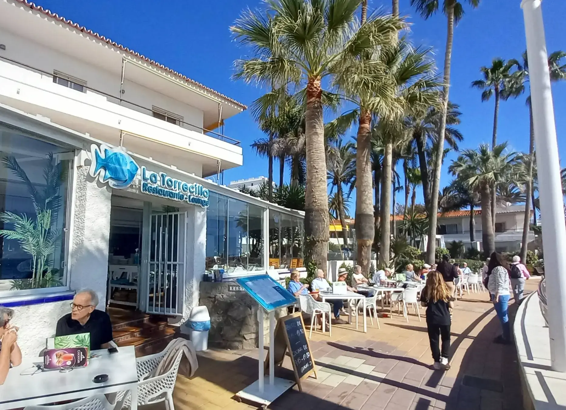 Fachada con el rótulo del pez azul y terraza bajo las palmeras del paseo