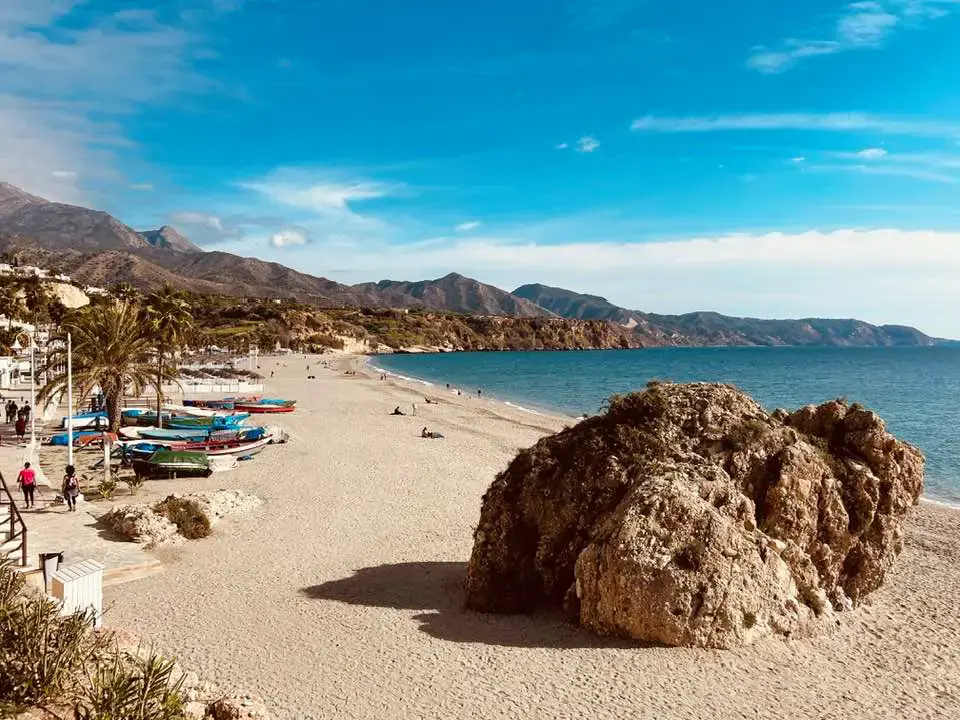 Gran roca en la arena de Burriana, con barcas varadas y las sierras de Nerja al fondo
