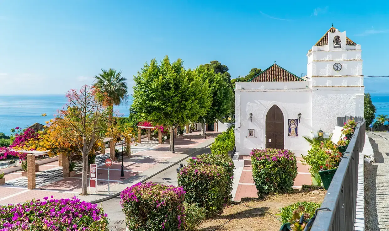 Iglesia blanca de Maro junto al paseo con buganvillas, palmeras y el mar detrás