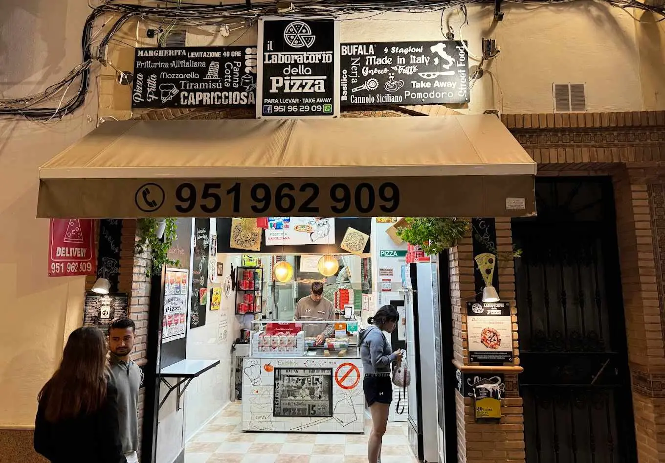 Local de Il laboratorio della pizza de noche, con mostrador para llevar y clientes en la puerta
