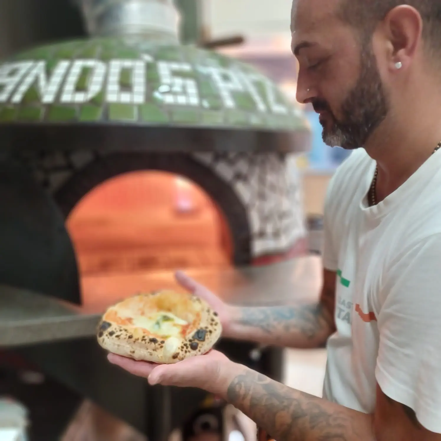 Pizzero mostrando una pizza recién hecha delante del horno de leña con mosaico verde