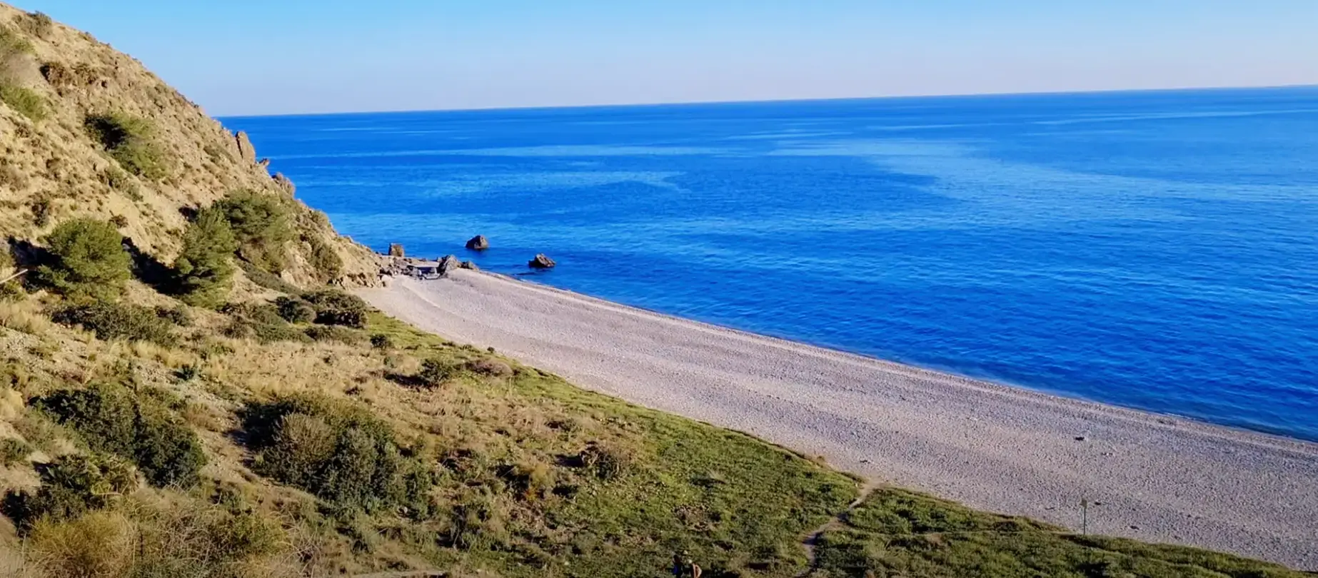 Larga orilla de grava de la playa de las Alberquillas bajo la ladera verde