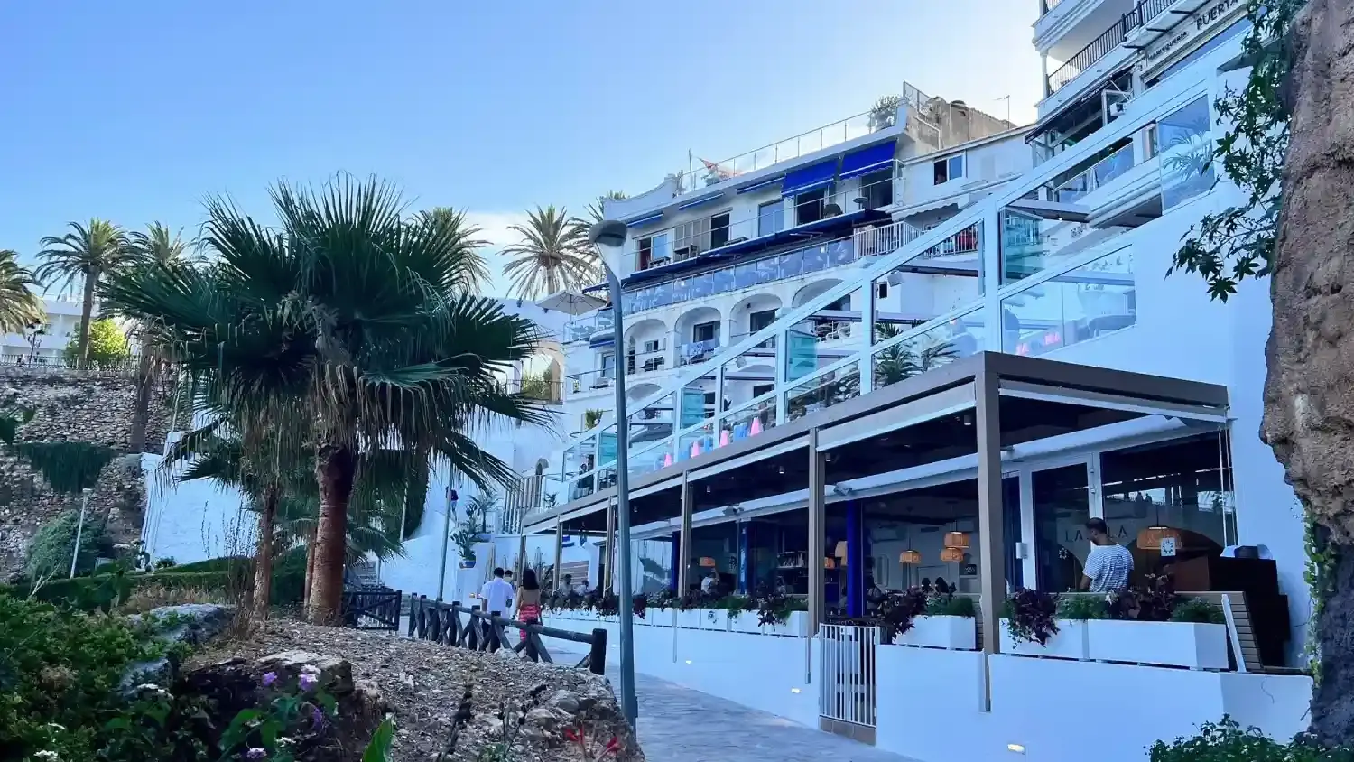 Terraza cubierta bajo los edificios blancos del acantilado, con palmeras y paseantes