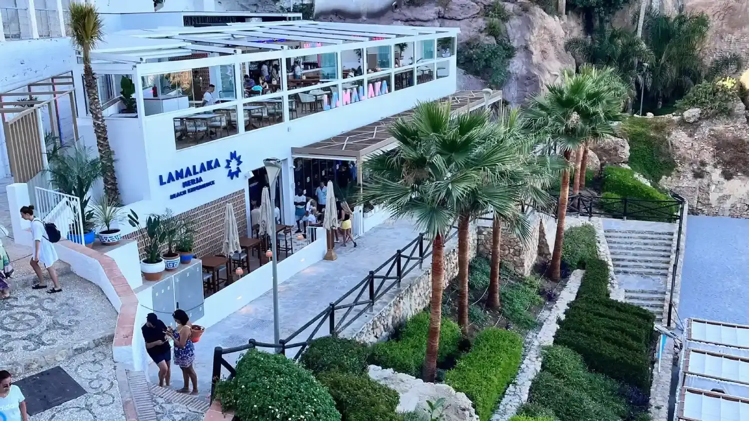 Edificio blanco del beach club con terraza acristalada, palmeras y escaleras a la playa