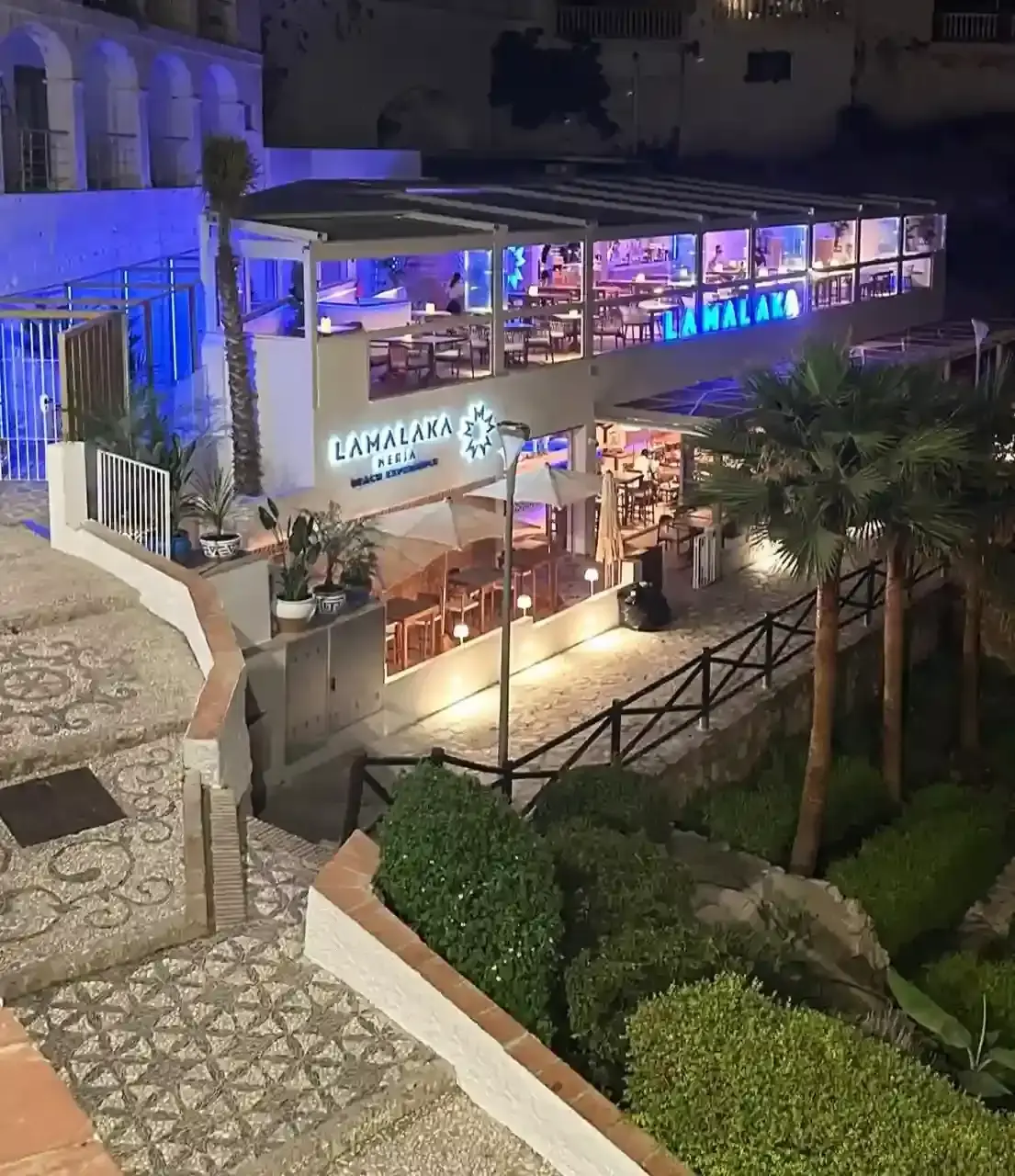 La Malaka de noche con luces azules, terraza acristalada y jardín con palmeras