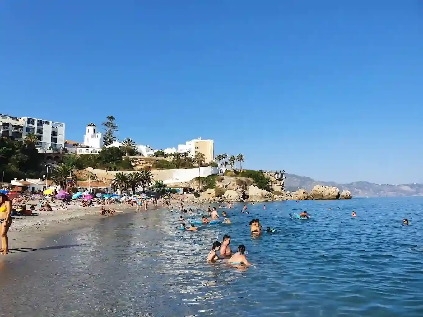 Bañistas en el agua clara de la playa El Salón, con las rocas y los edificios del acantilado
