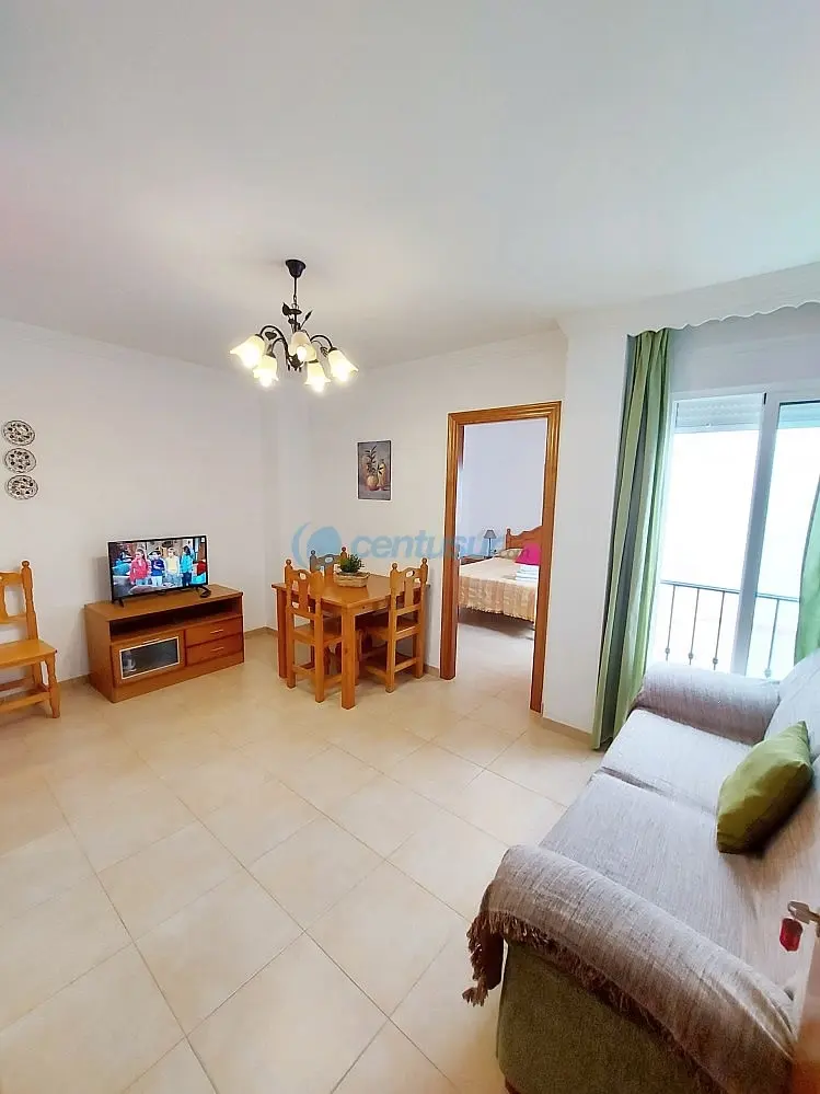 Salón con sofá, mesa de comedor de madera, televisión y balcón, con el dormitorio al fondo
