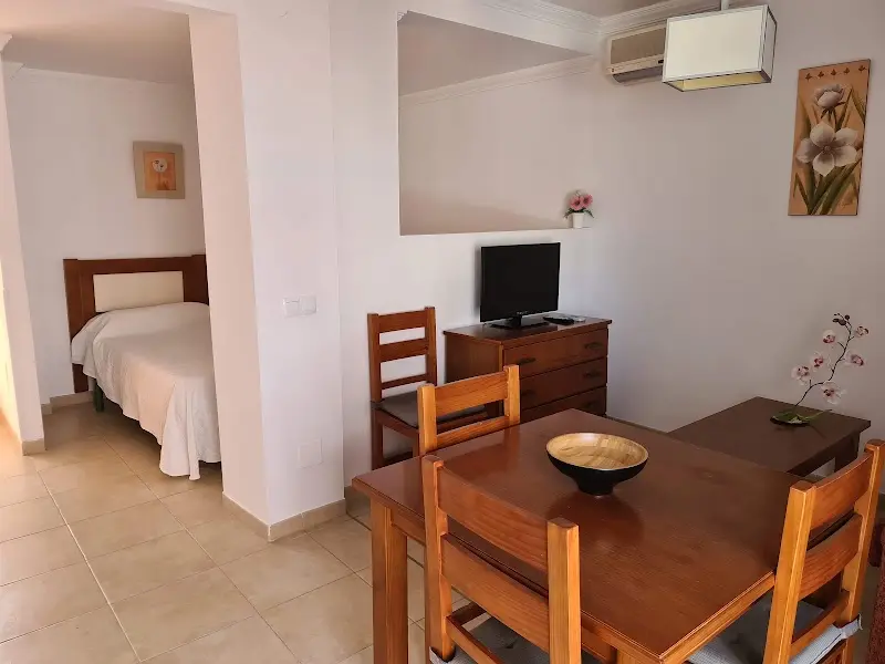 Salón comedor con mesa de madera y televisión, con un dormitorio de cama individual al lado