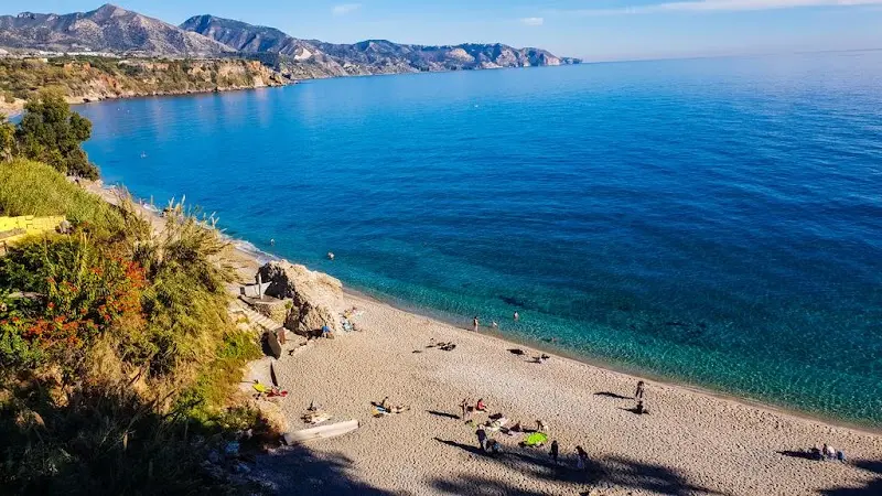 Playa de arena con bañistas y agua turquesa vista desde lo alto, con la sierra al fondo