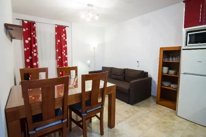 Salón comedor con mesa de madera, sofá marrón, cortinas rojas y nevera junto a la cocina