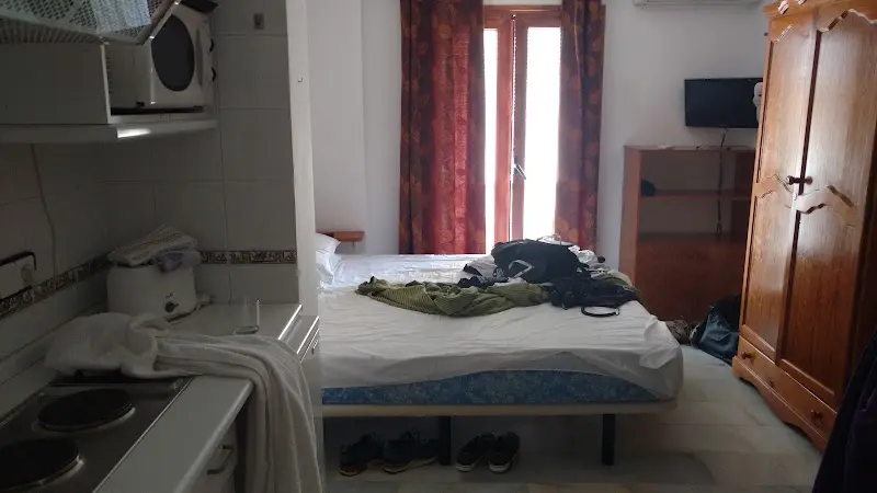 Estudio con cama de matrimonio, cocina pequeña, armario de madera y balcón con cortinas