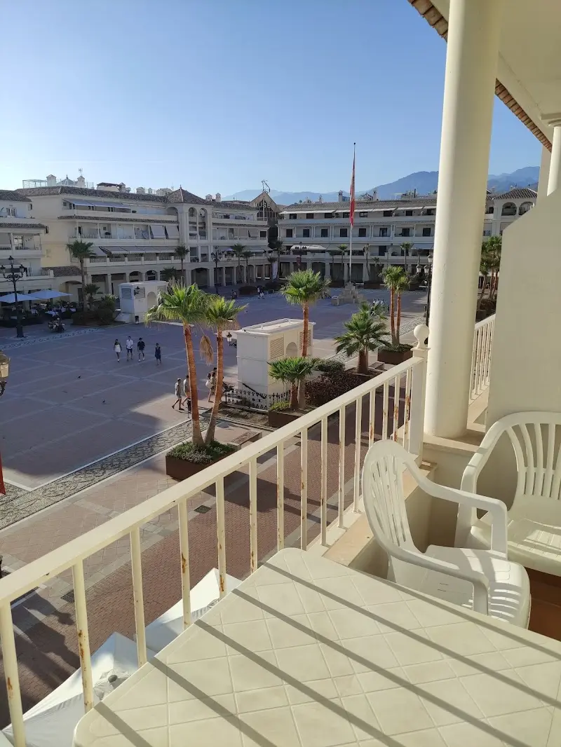 Balcón con sillas blancas y vistas a una gran plaza con palmeras y las montañas