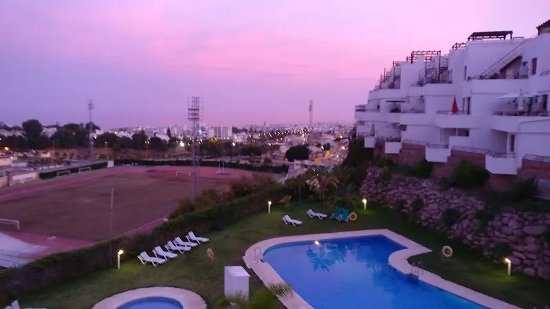 Piscinas y jardín iluminados al anochecer bajo un cielo rosado, junto a los apartamentos