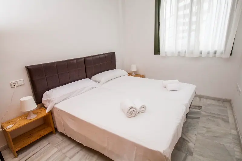 Dormitorio con dos camas juntas, cabecero de piel oscura, toallas y mesillas de madera
