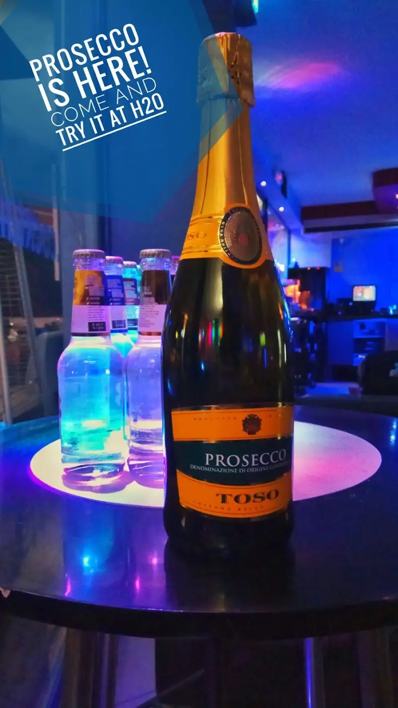 Botella de prosecco sobre una mesa iluminada con el anuncio «Prosecco is here! Come and try it at H2O»