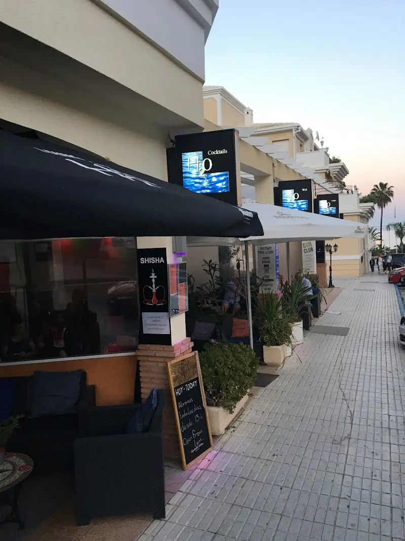Terraza del Bar H2O al atardecer, con toldo negro, sofás, pizarra y carteles luminosos de cócteles
