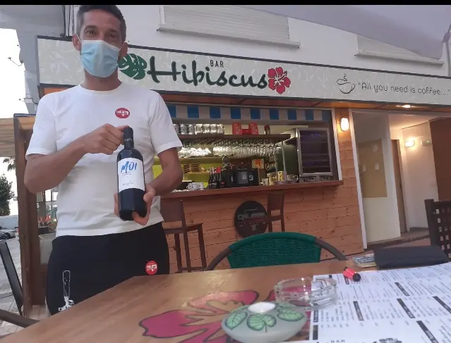 Camarero mostrando una botella de vino tinto en la terraza, con la barra del bar detrás