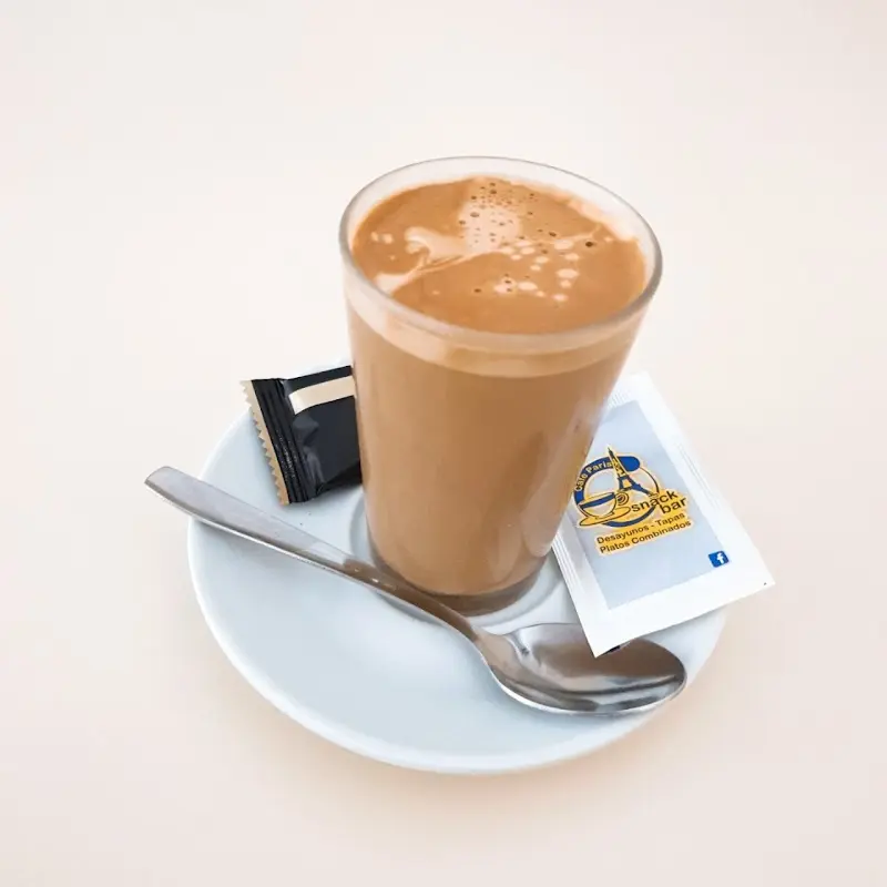 Café con leche en vaso sobre platito, con cucharilla y sobre de azúcar de Cafe Paris