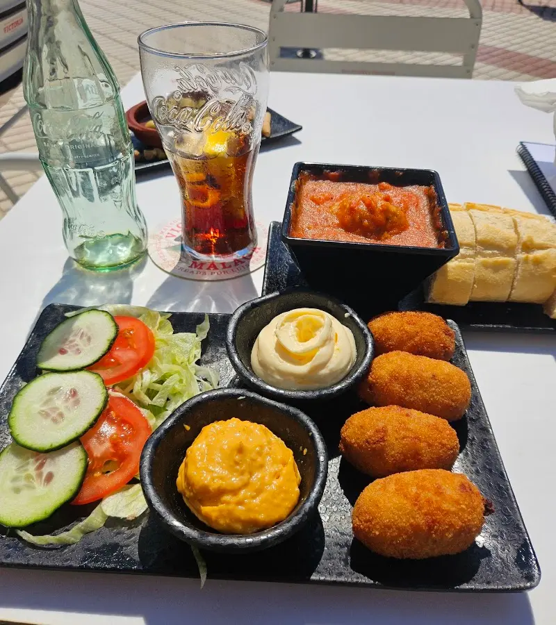 Croquetas, salsas, pan y un refresco en una mesa al sol de The Corner
