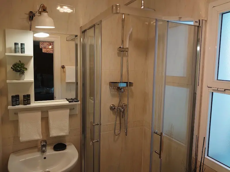 Baño con ducha de mampara, lavabo, estantes con amenities y toallas blancas