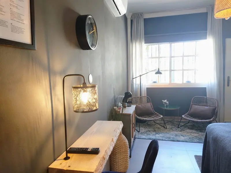 Rincón de la habitación con escritorio de madera, lámpara, reloj y dos sillones junto a la ventana