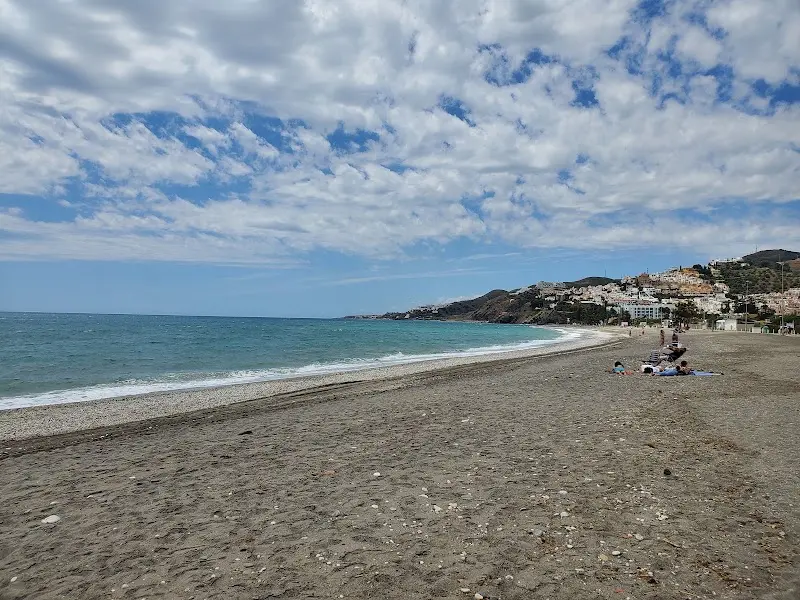 Arena oscura de El Playazo con nubes y Nerja al fondo sobre los acantilados